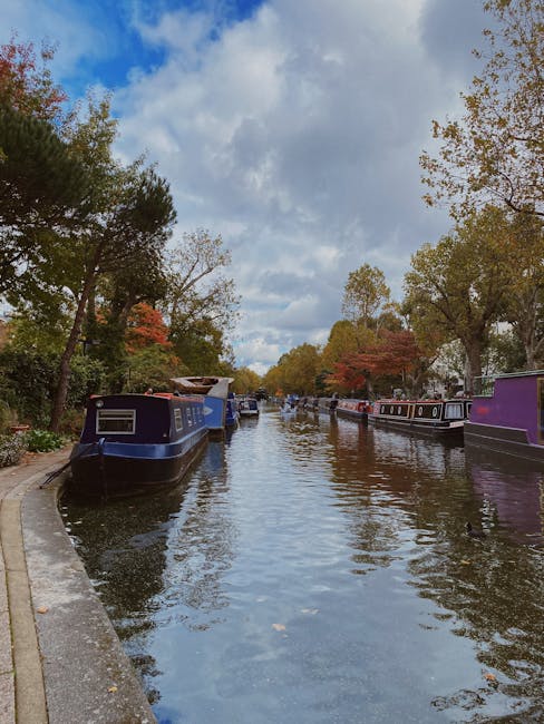 A tranquil canal-side scene in Little Venice, with lush green trees lining both sides of the water. Several small pedal boats with bright red, blue, and pink hues are occupied by people enjoying leisu